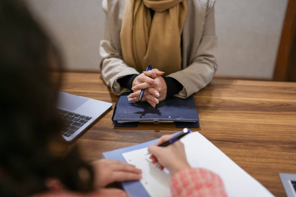 Job interview in progress at a conference table, with a candidate holding a pen and listening while another person takes notes. A laptop and folders are visible on the table, creating a professional office setting.