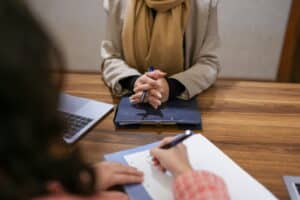 Job interview in progress at a conference table, with a candidate holding a pen and listening while another person takes notes. A laptop and folders are visible on the table, creating a professional office setting.