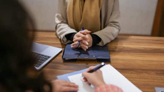 Job interview in progress at a conference table, with a candidate holding a pen and listening while another person takes notes. A laptop and folders are visible on the table, creating a professional office setting.