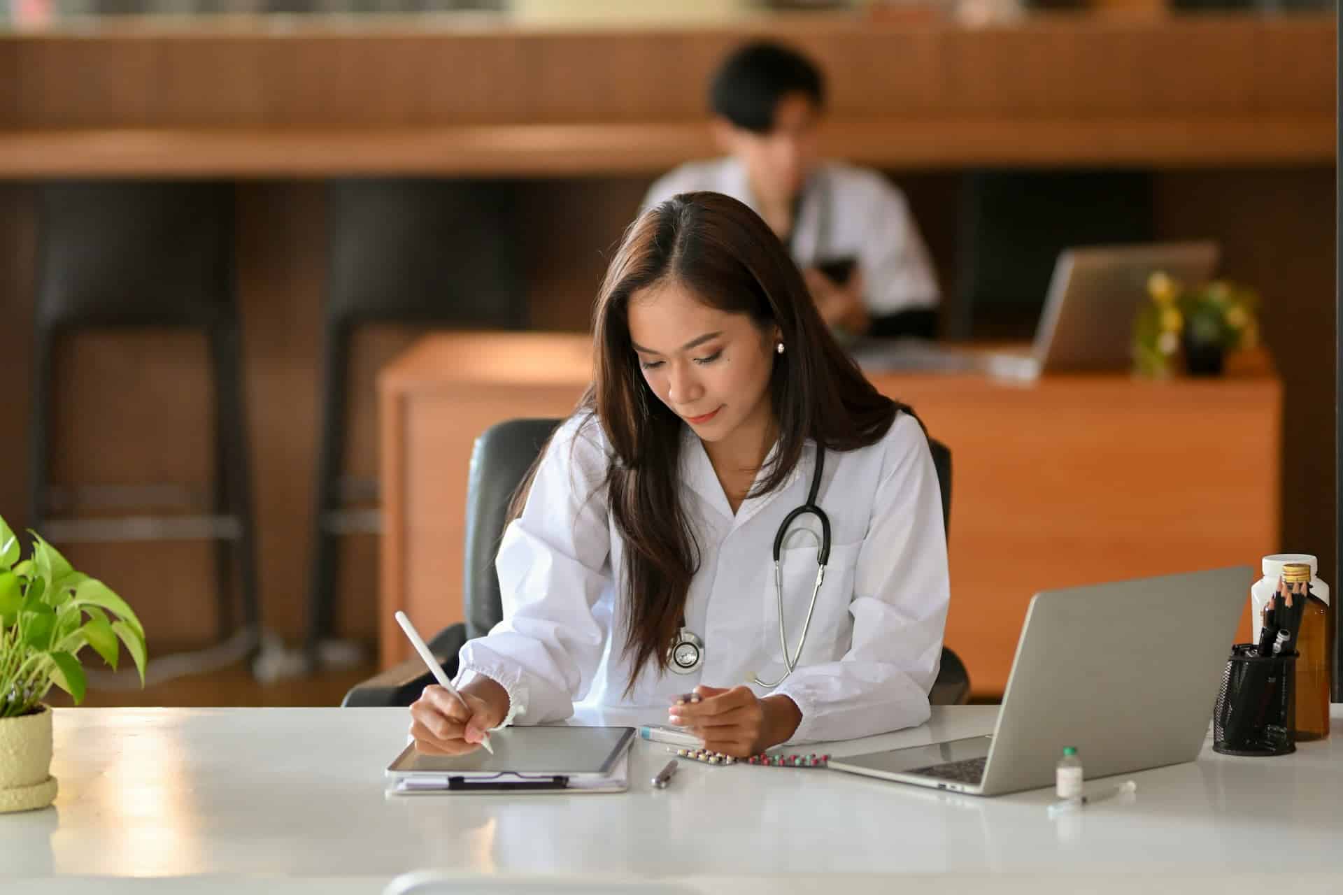 Medical professional in white coat with stethoscope writing notes at a desk with a laptop and medication in a clinical office setting