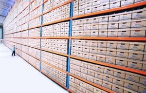 Large warehouse archive with tall metal shelving units filled with neatly stacked labeled document storage boxes, with a person walking down the aisle for scale.