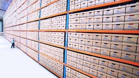 Large warehouse archive with tall metal shelving units filled with neatly stacked labeled document storage boxes, with a person walking down the aisle for scale.