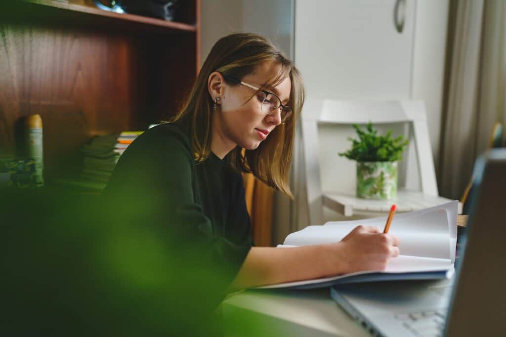 Woman wearing glasses reviewing and editing a document at a desk with a laptop and notebook.
