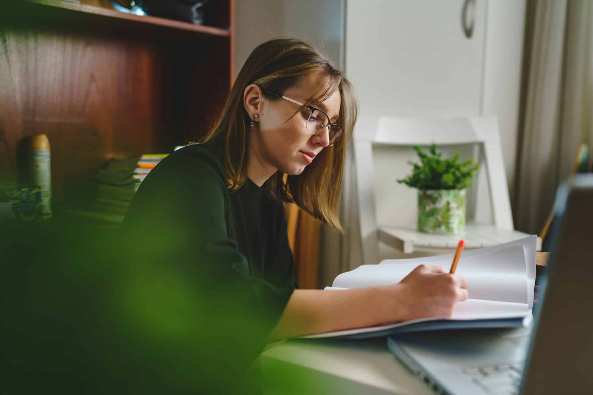 Woman wearing glasses reviewing and editing a document at a desk with a laptop and notebook.