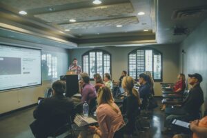 Instructor giving a presentation at the front of a small classroom while adult learners sit in desks watching a projected slide.