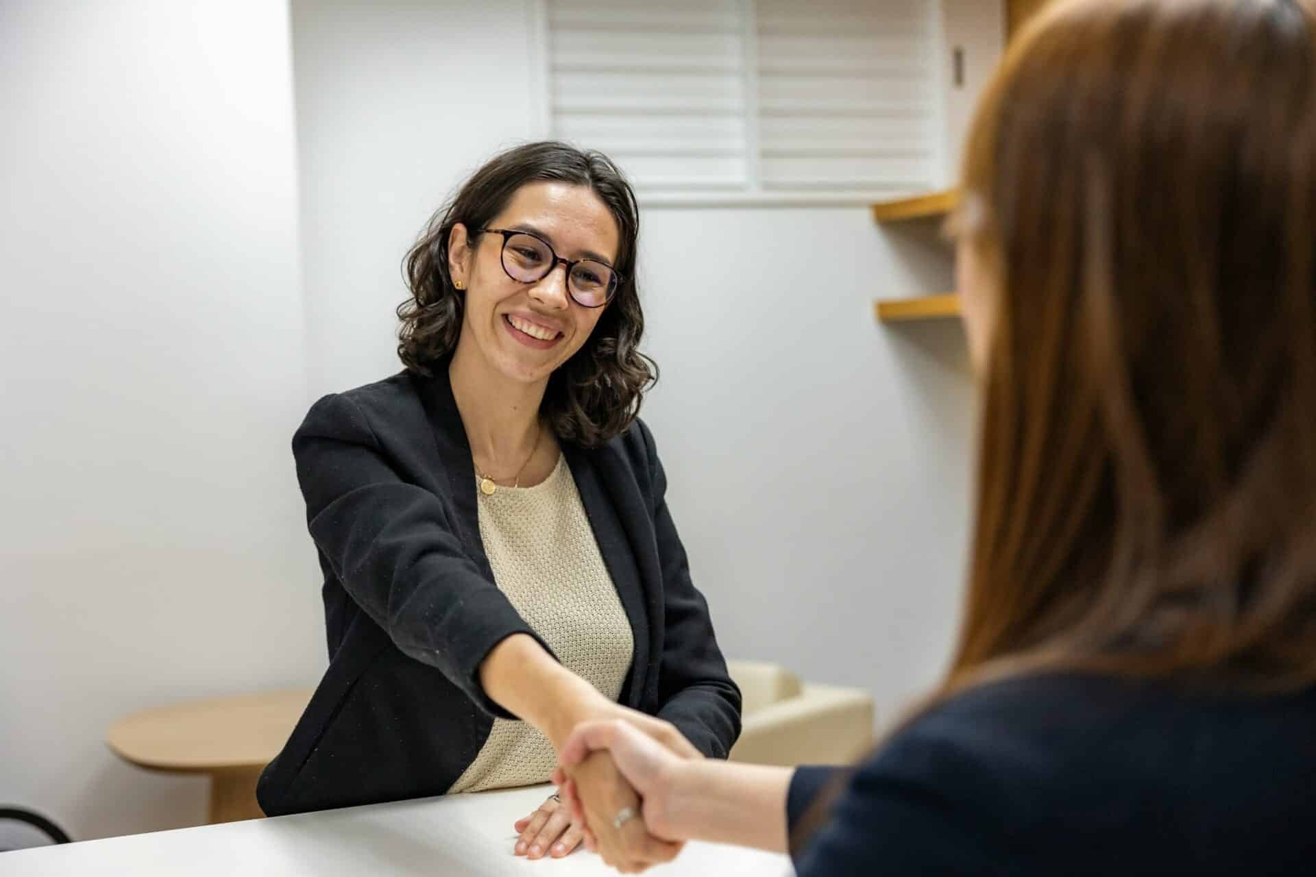 Two professionals shaking hands across a desk during a meeting or interview in an office setting.