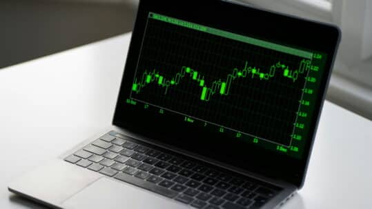 Laptop displaying a green candlestick stock chart on a dark screen, showing market price fluctuations over time.