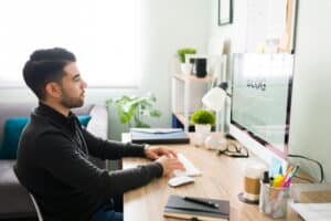 Man writing a blog post at a desk, working on a desktop computer in a bright home office with plants and office supplies nearby.