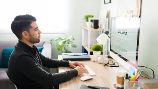 Man writing a blog post at a desk, working on a desktop computer in a bright home office with plants and office supplies nearby.