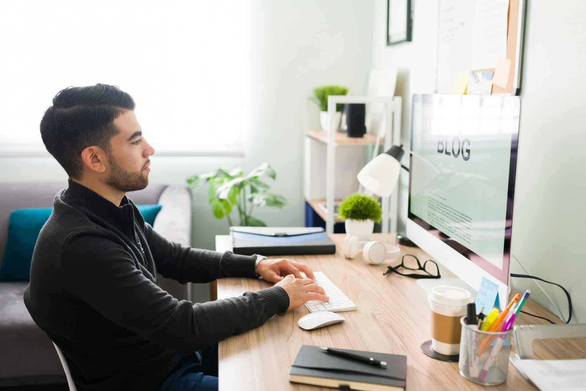 Man writing a blog post at a desk, working on a desktop computer in a bright home office with plants and office supplies nearby.