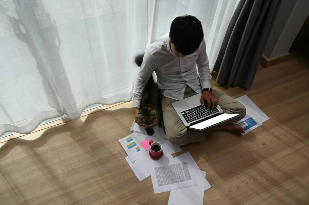 Person sitting cross-legged on a wooden floor using a laptop, reviewing printed charts and documents, with a cat beside them and a cup of coffee nearby.