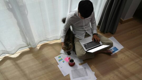 Person sitting cross-legged on a wooden floor using a laptop, reviewing printed charts and documents, with a cat beside them and a cup of coffee nearby.