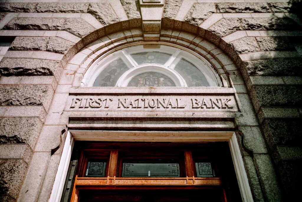Stone entrance of a historic bank building with “First National Bank” engraved above an arched window and wooden doorway.