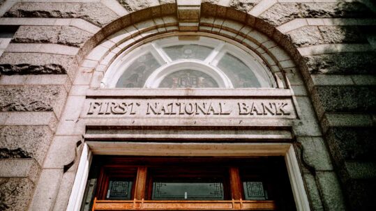 Stone entrance of a historic bank building with “First National Bank” engraved above an arched window and wooden doorway.