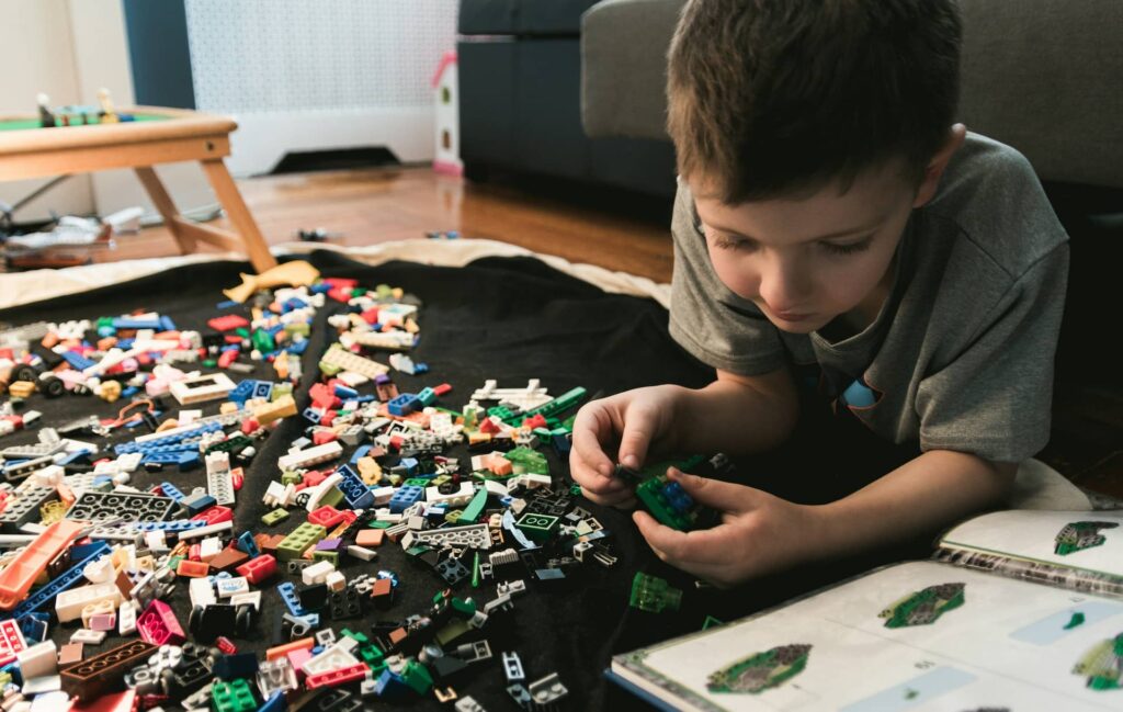 A young child lying on the floor assembling a small LEGO build, surrounded by scattered colorful bricks and an open instruction booklet.