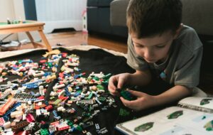 A young child lying on the floor assembling a small LEGO build, surrounded by scattered colorful bricks and an open instruction booklet.