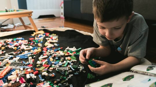 A young child lying on the floor assembling a small LEGO build, surrounded by scattered colorful bricks and an open instruction booklet.