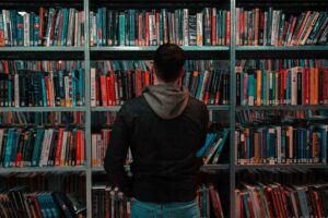 Person standing in front of a bookshelf filled with books, viewed from behind, appearing to browse or choose a book in a library or bookstore.
