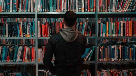 Person standing in front of a bookshelf filled with books, viewed from behind, appearing to browse or choose a book in a library or bookstore.