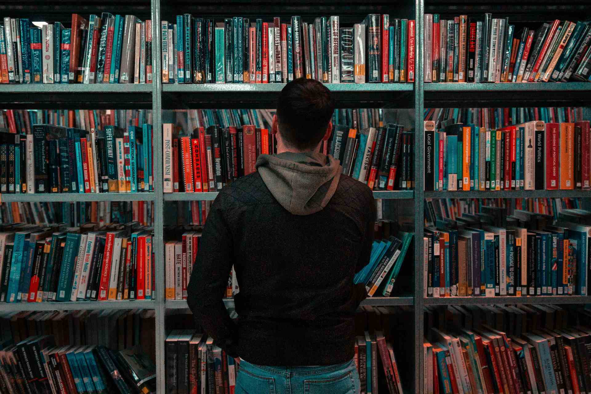 Person standing in front of a bookshelf filled with books, viewed from behind, appearing to browse or choose a book in a library or bookstore.