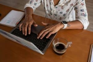 Person typing on a laptop at a wooden desk with a notebook and a cup of coffee nearby, wearing a smartwatch.