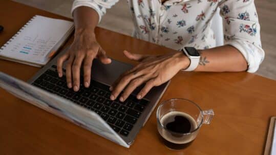Person typing on a laptop at a wooden desk with a notebook and a cup of coffee nearby, wearing a smartwatch.
