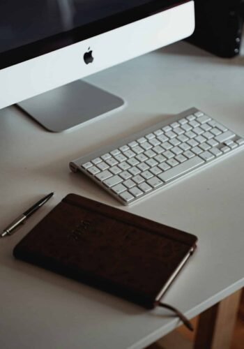 A clean workspace featuring an Apple desktop computer, wireless keyboard, pen, and a closed leather notebook on a white desk.