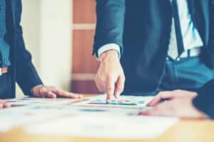 Close-up of business professionals in suits reviewing documents on a table, with one person pointing at a chart or color-coded graphics during a meeting.