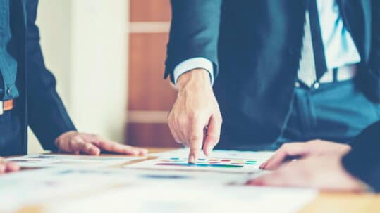 Close-up of business professionals in suits reviewing documents on a table, with one person pointing at a chart or color-coded graphics during a meeting.