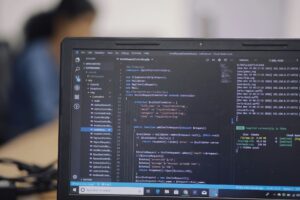 Close-up of a laptop screen displaying code in a development environment, with a blurred person working in the background.