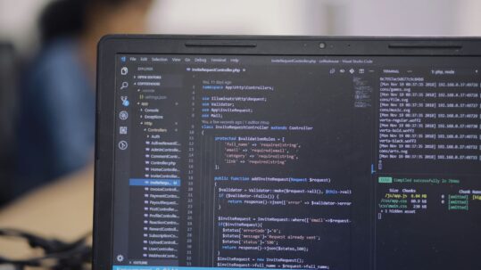 Close-up of a laptop screen displaying code in a development environment, with a blurred person working in the background.