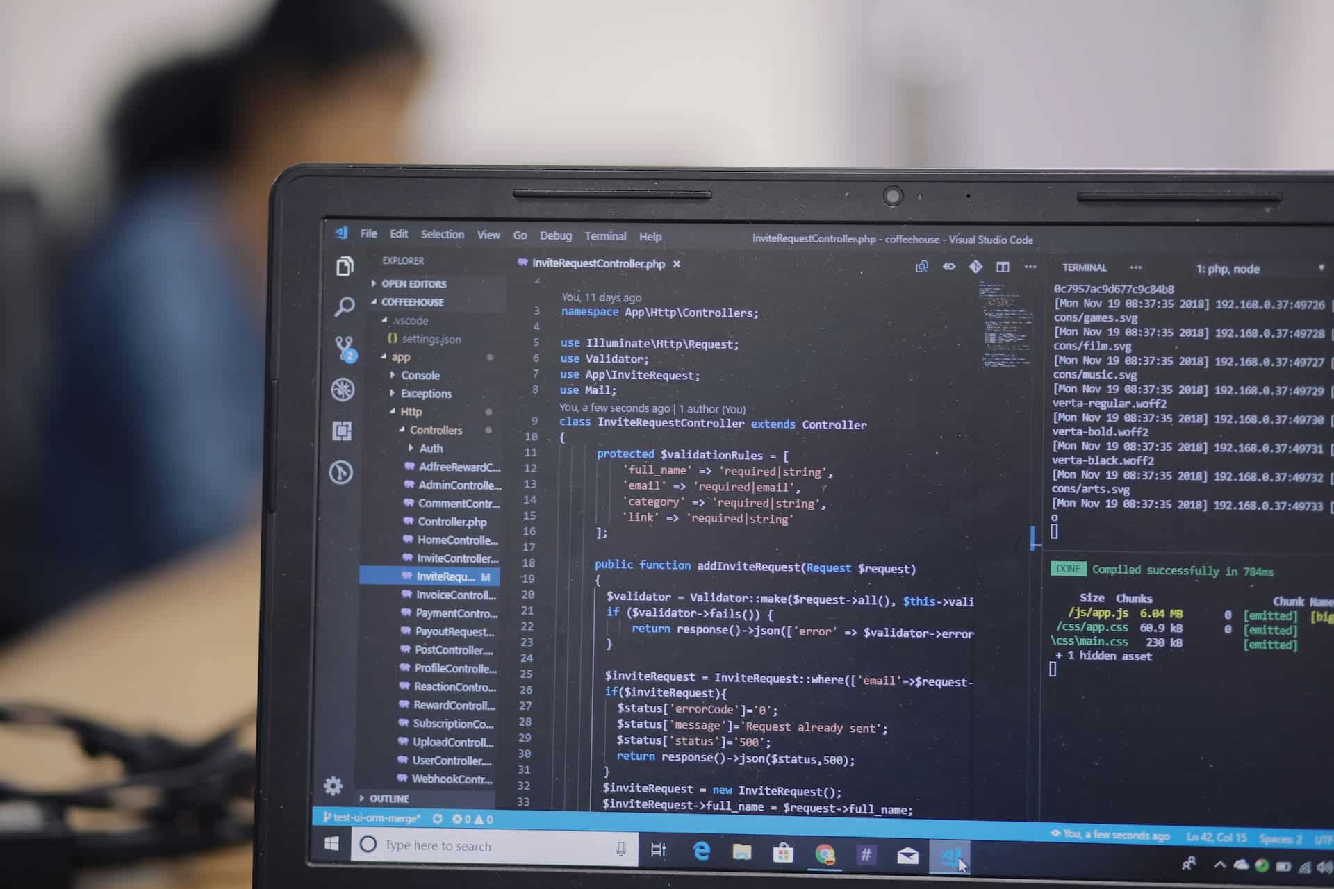 Close-up of a laptop screen displaying code in a development environment, with a blurred person working in the background.