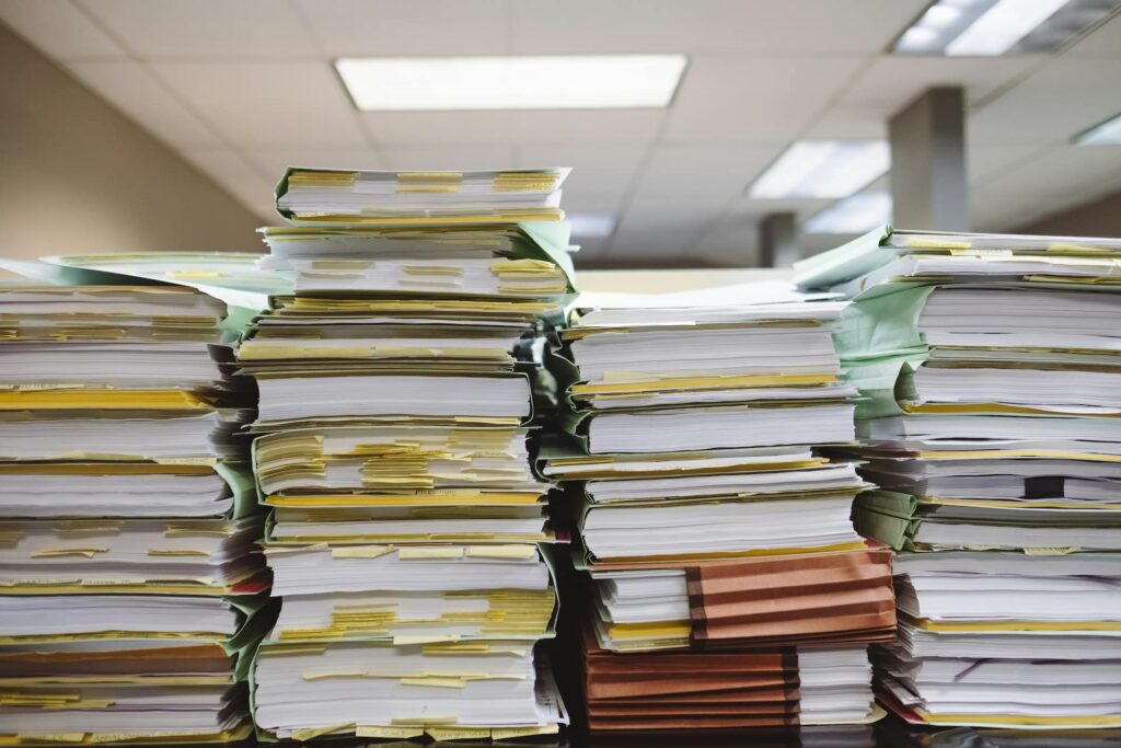 Stacks of thick paper files and binders piled high on office desks, covered with sticky notes and tabs, in a fluorescent-lit workspace, conveying a heavy administrative workload and document overload.