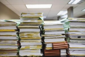 Stacks of thick paper files and binders piled high on office desks, covered with sticky notes and tabs, in a fluorescent-lit workspace, conveying a heavy administrative workload and document overload.