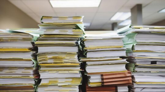 Stacks of thick paper files and binders piled high on office desks, covered with sticky notes and tabs, in a fluorescent-lit workspace, conveying a heavy administrative workload and document overload.