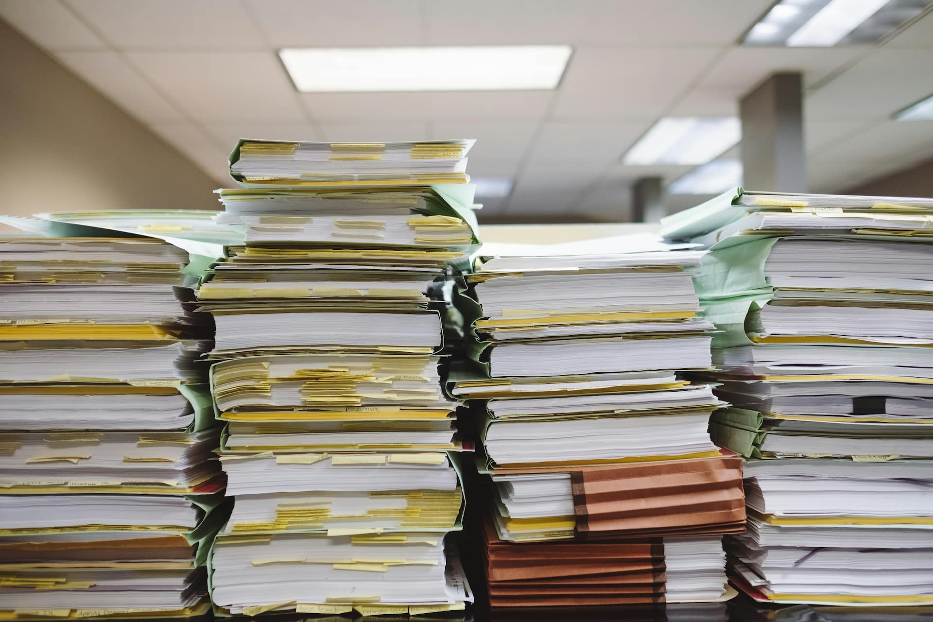 Stacks of thick paper files and binders piled high on office desks, covered with sticky notes and tabs, in a fluorescent-lit workspace, conveying a heavy administrative workload and document overload.