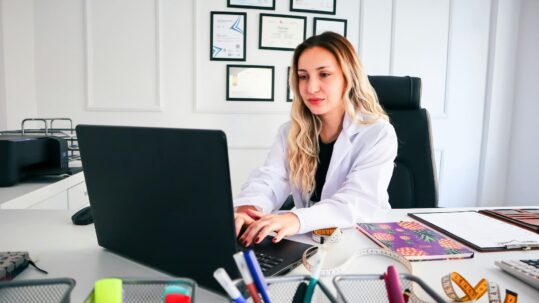 Healthcare professional reviewing documentation on a laptop in a medical office, illustrating healthcare technical writing, compliance, and documentation management.