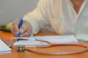 Healthcare professional writing notes on a document at a desk, with a stethoscope in the foreground and paperwork spread across the table. The image focuses on the hand holding a blue pen, symbolizing medical documentation and patient recordkeeping.