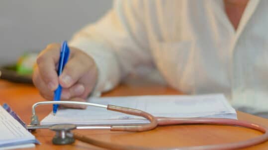 Healthcare professional writing notes on a document at a desk, with a stethoscope in the foreground and paperwork spread across the table. The image focuses on the hand holding a blue pen, symbolizing medical documentation and patient recordkeeping.