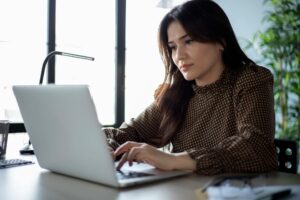 Woman working on a laptop at a desk in a bright office, focused on typing, with a desk lamp and plants in the background.