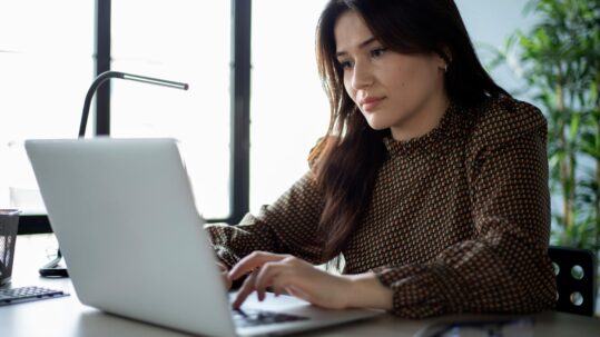 Woman working on a laptop at a desk in a bright office, focused on typing, with a desk lamp and plants in the background.