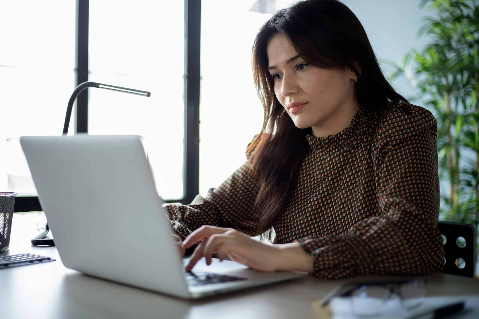 Woman working on a laptop at a desk in a bright office, focused on typing, with a desk lamp and plants in the background.