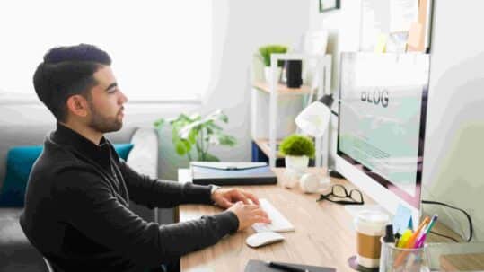 Marketing technical writer creating a blog post on a desktop computer in a modern office workspace with a notebook, coffee, and office supplies.