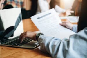 Person reviewing a printed document while working on a laptop at a conference table, with colleagues collaborating in the background.