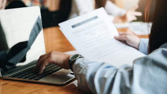 Person reviewing a printed document while working on a laptop at a conference table, with colleagues collaborating in the background.