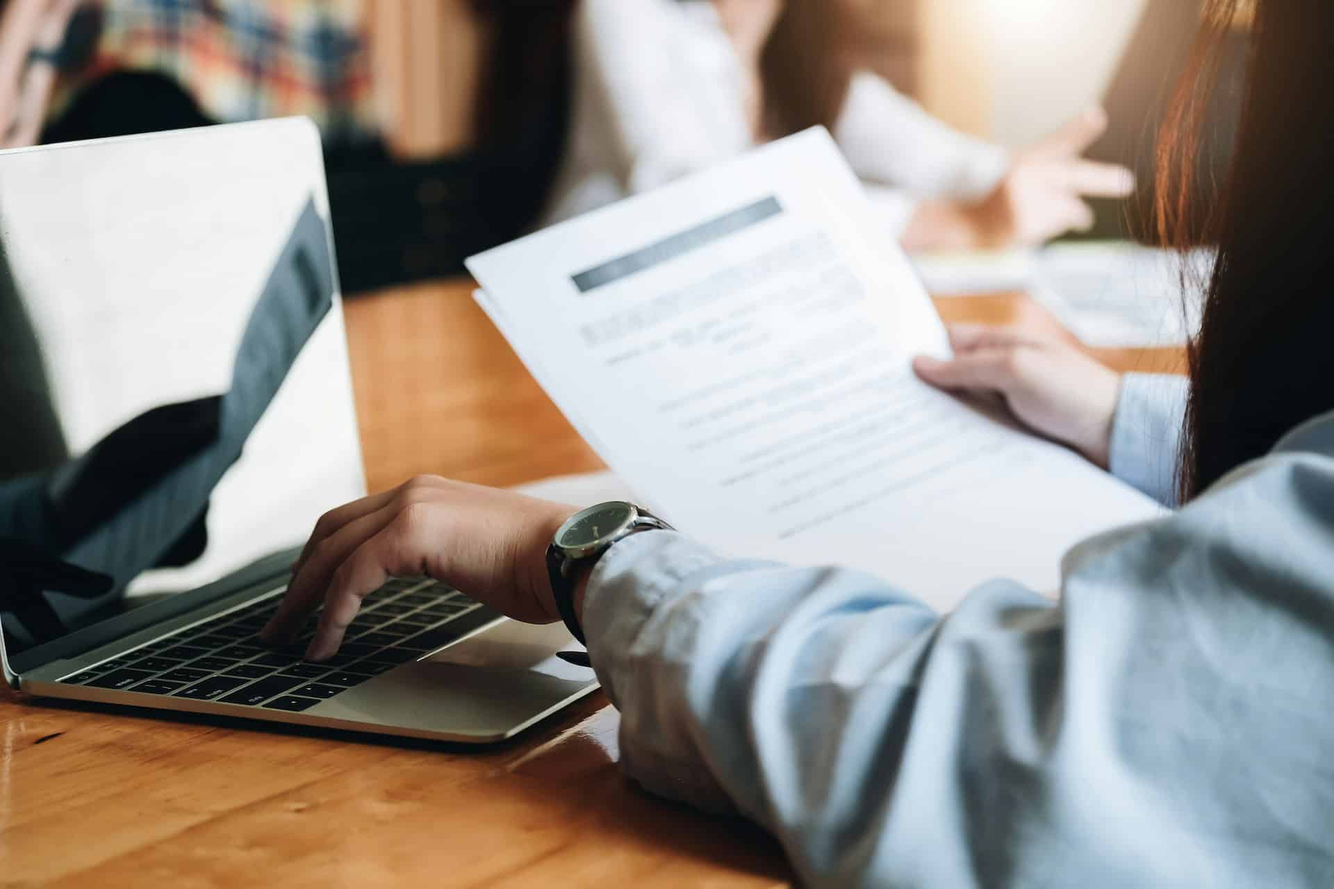 Person reviewing a printed document while working on a laptop at a conference table, with colleagues collaborating in the background.