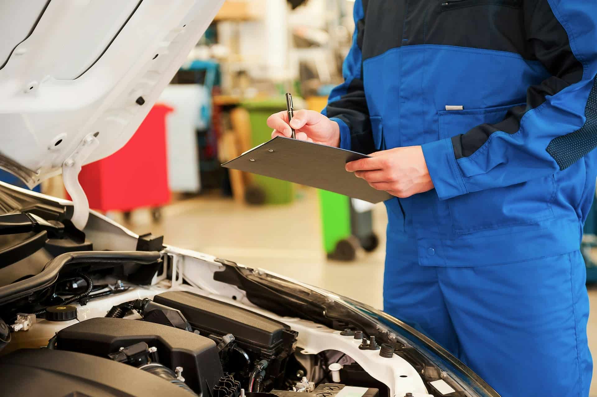 Automotive technician inspecting a vehicle engine while documenting service procedures on a clipboard.