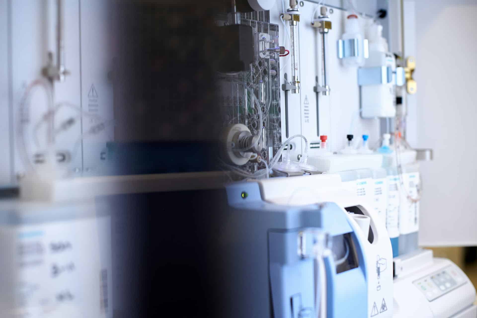Close-up of medical device equipment and monitoring systems in a clean healthcare or laboratory environment, with tubing, control panels, and diagnostic instruments visible.