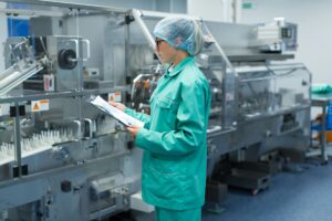 Pharmaceutical manufacturing professional reviewing documentation on a clipboard beside production equipment.