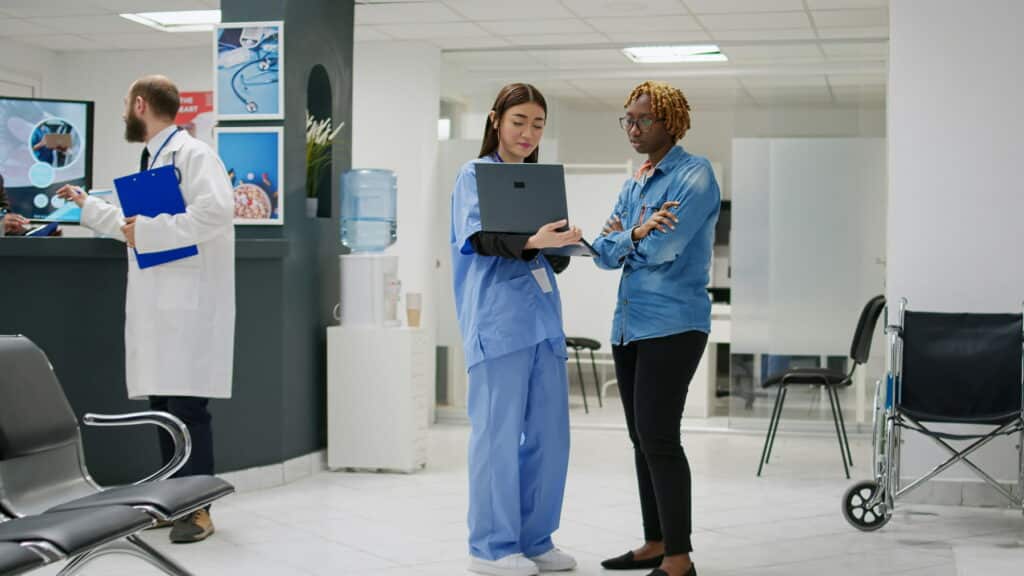 Healthcare professional in blue scrubs reviewing a laptop with another person in a medical clinic waiting area.